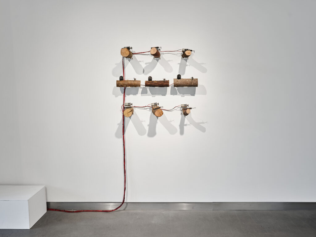 View of sound artwork on gallery wall. The wall is a 3x3 grid of 9 wooden logs, DC motors and trip alarms with long red and black electrical cables zip-tied together coming down to floor. Electrical cable moves left along floor into a white box.