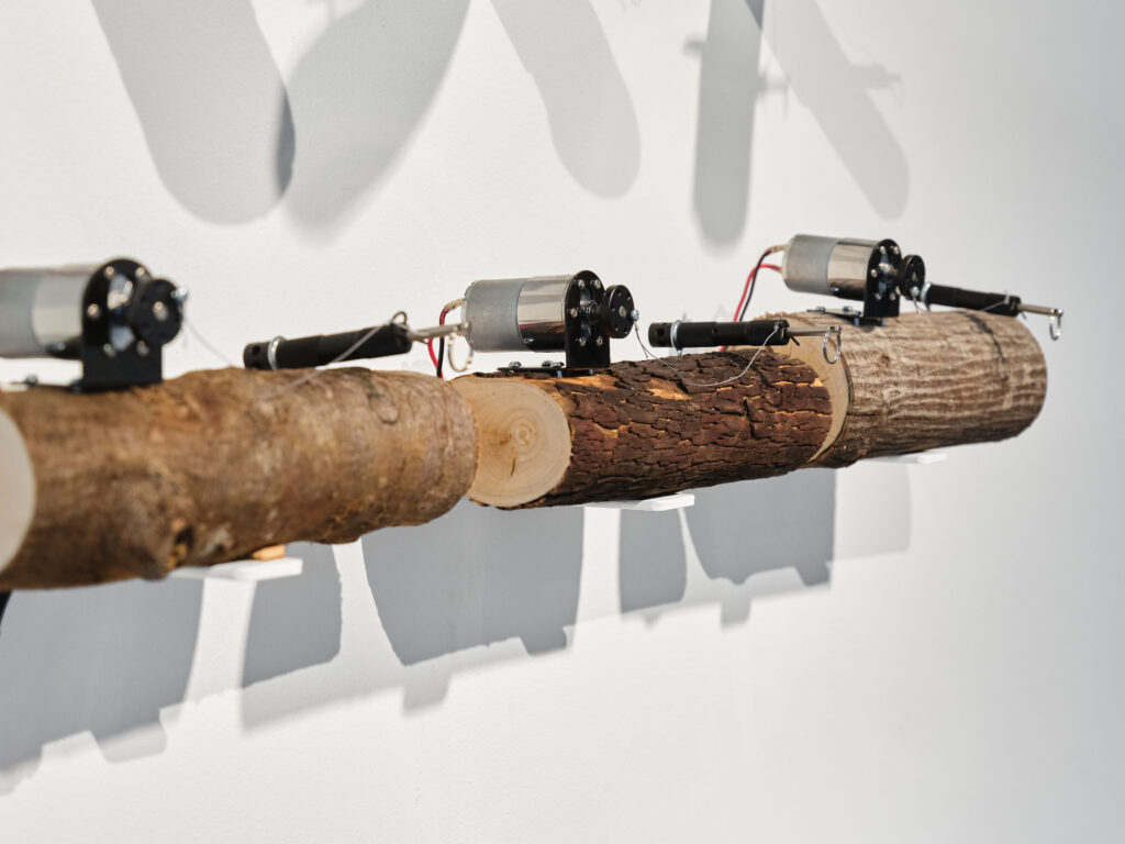 Side view of three 10" logs against gallery wall. DC motors sit on top left of logs with perimeter trip alarms on top right of logs. The view shows 1/3 of sound artwork, S.O.S.