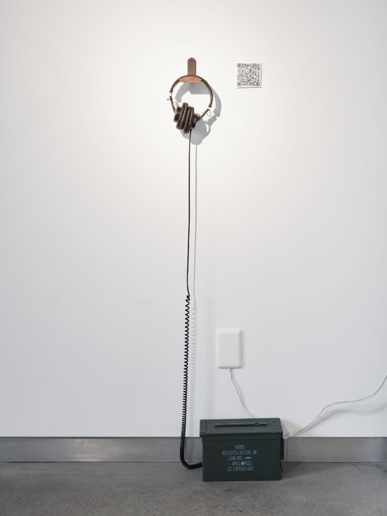 A pair of headphones rest on gallery wall with cord disappearing inside a 50 caliber ammo box resting on the floor.