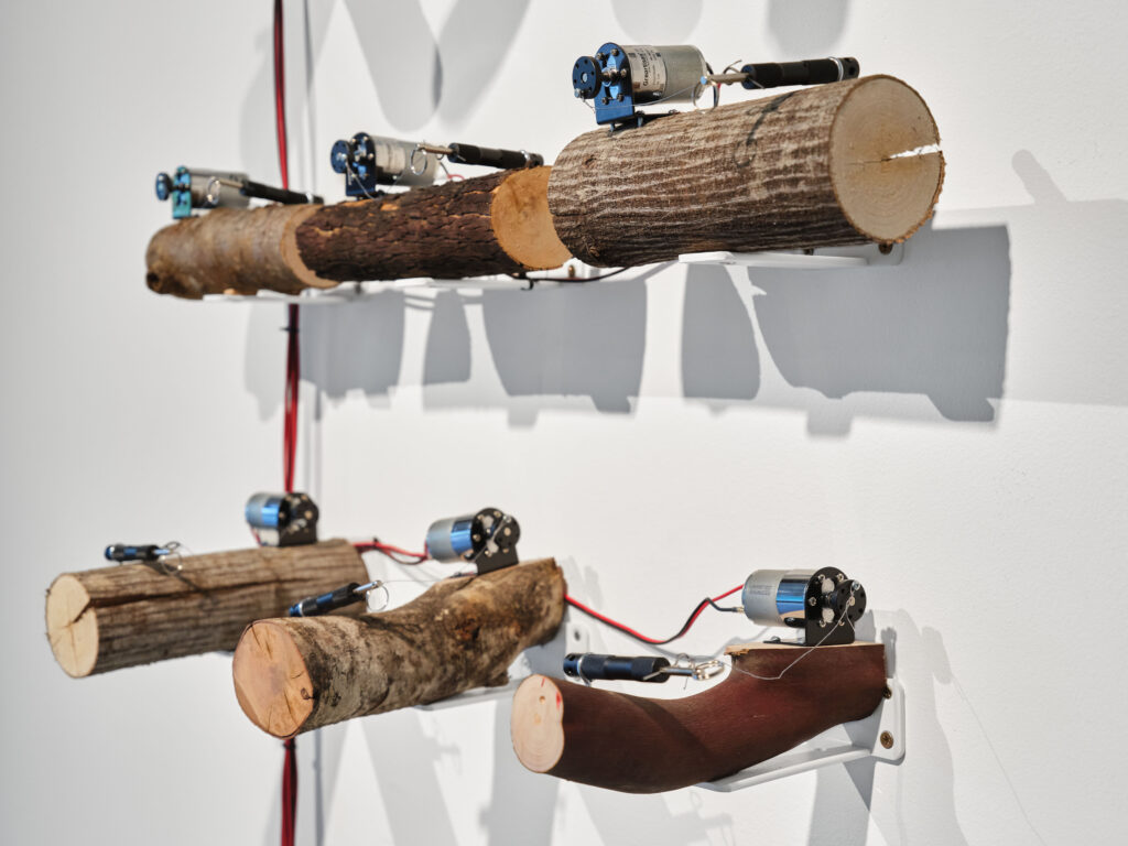 Right side view of six 10" logs against gallery wall. DC motors sit on top left of logs with perimeter trip alarms on top right of logs. The view shows 2/3 of sound artwork, S.O.S.