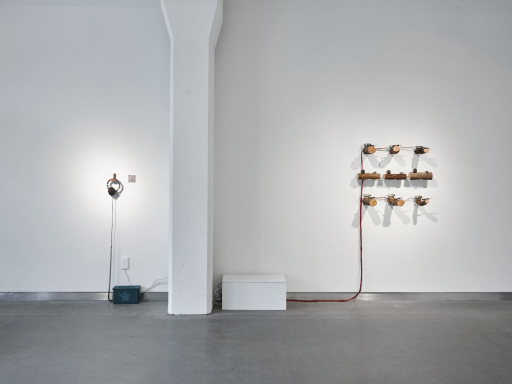 distant view of two artworks on gallery wall with lighting center on each artwork. On left is a pair of headphones resting on wall that is connected to 50-caliber ammo box on floor. On right are 9 wooden logs, DC motors and trip alarms with long red and black electrical cables zip-tied together coming down to floor and into a box in middle of floor.